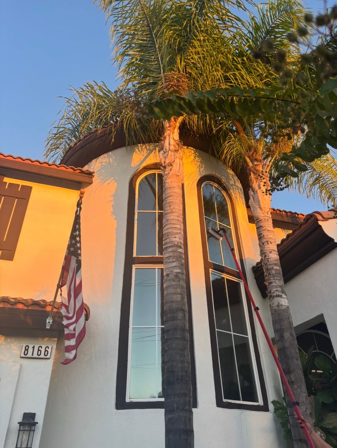 Long-handled brush cleaning tall arched windows on a house with palm trees and American flag.