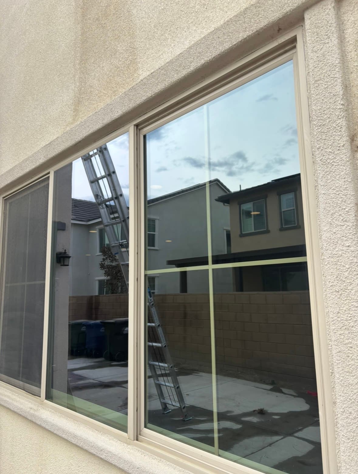 Large window in a stucco wall reflecting a ladder, brick wall, and neighboring houses.