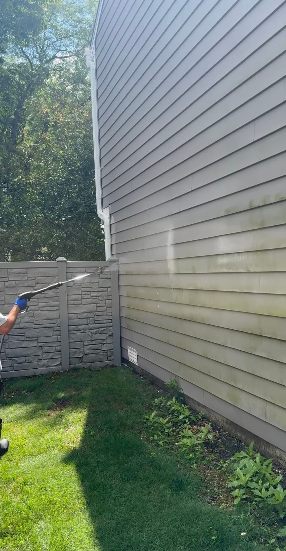 A person uses a pressure washer to clean green algae from grey vinyl house siding.