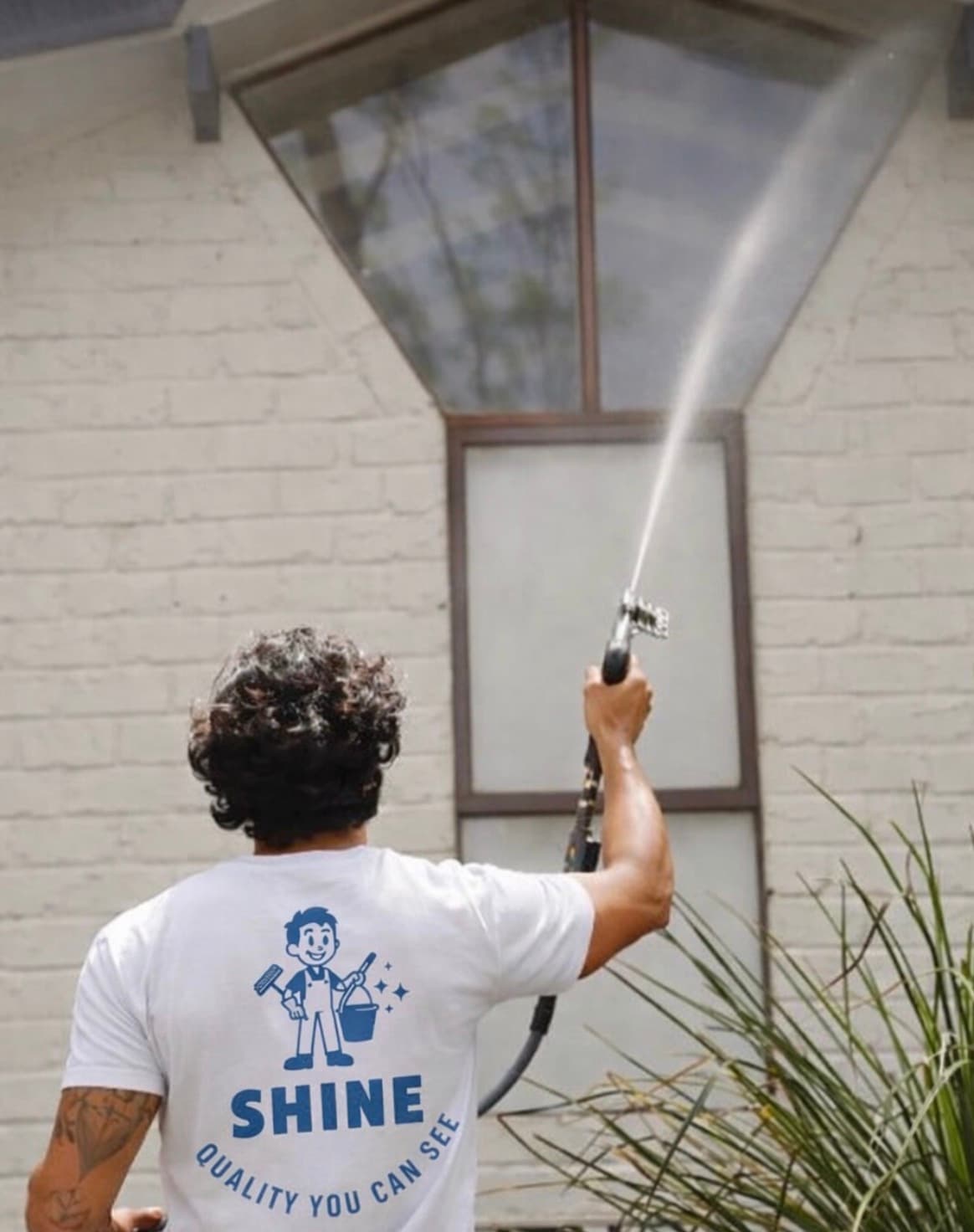 Man in a Shine t-shirt pressure washing a high window on a white brick wall.