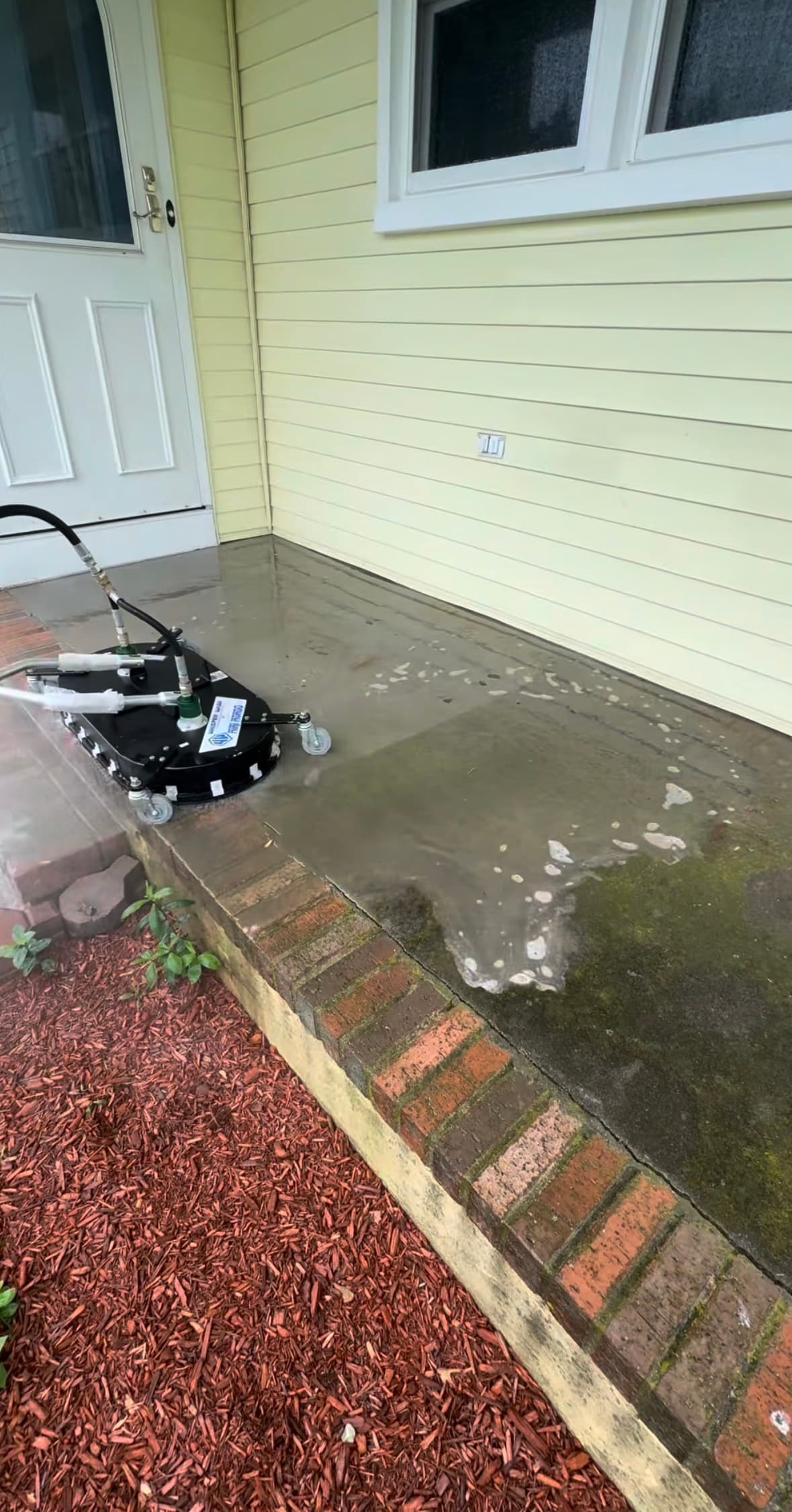 A surface cleaner removes moss and grime from a concrete porch with brick edging.